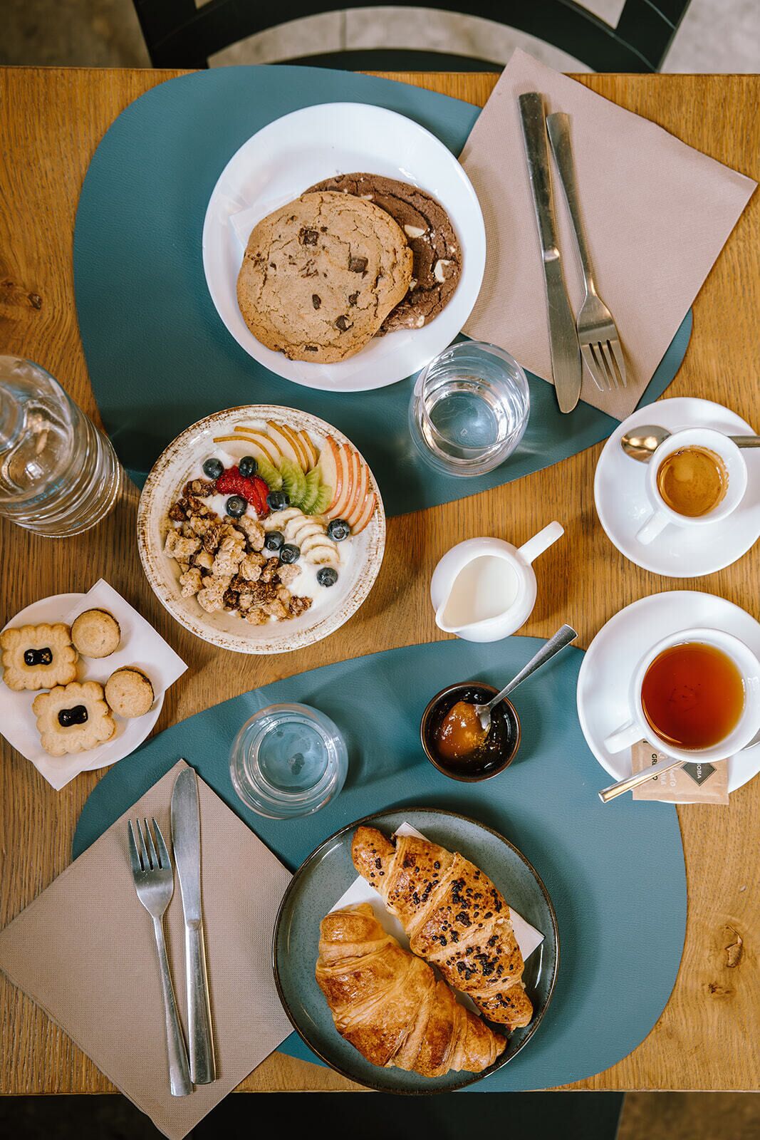 A breakfast setting with a plate of cookies, a bowl of cereal with fruit, a croissant, and a cup of tea.