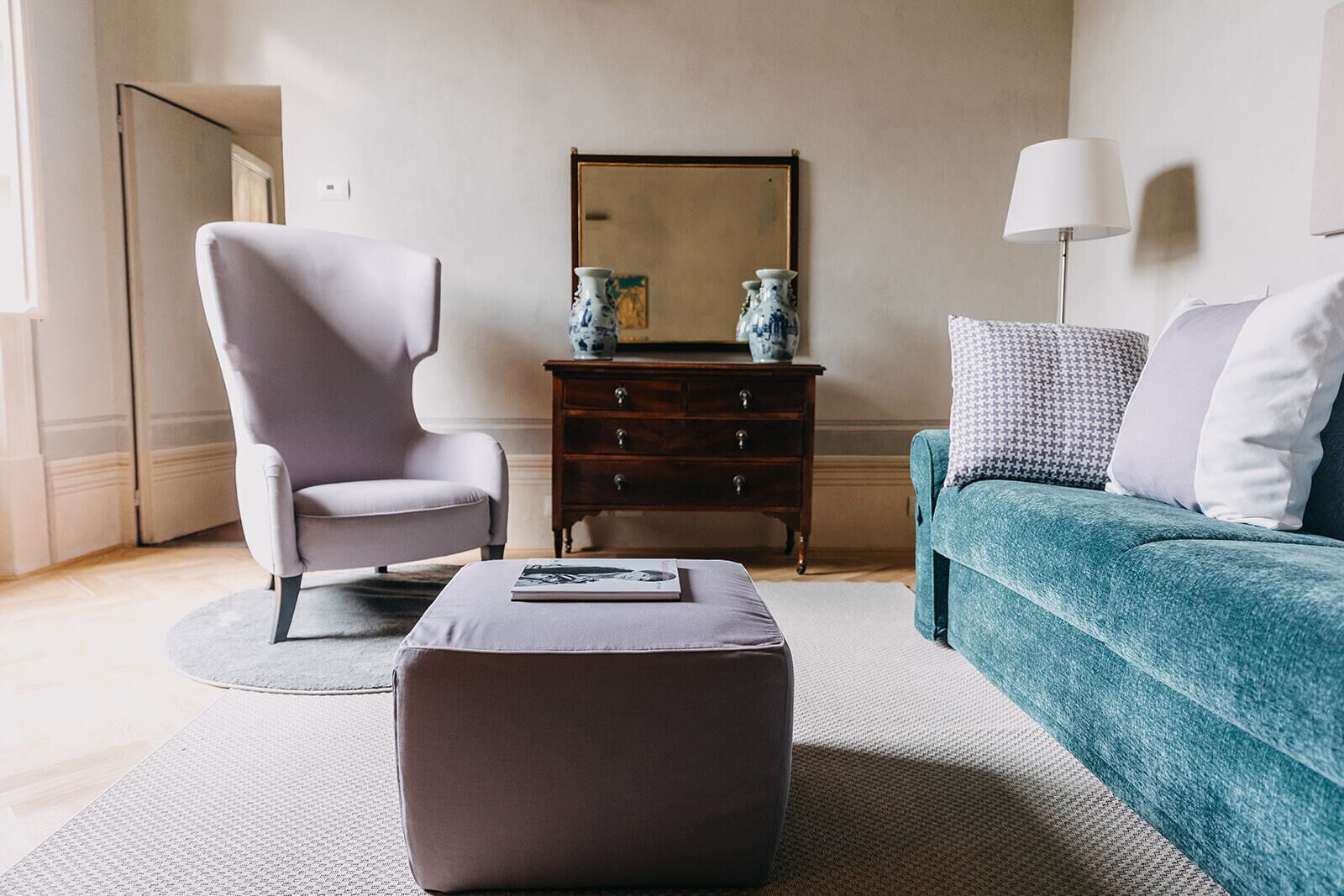 A modern living room with a grey armchair, a teal sofa, a wooden side table with decorative vases, and a floor lamp.