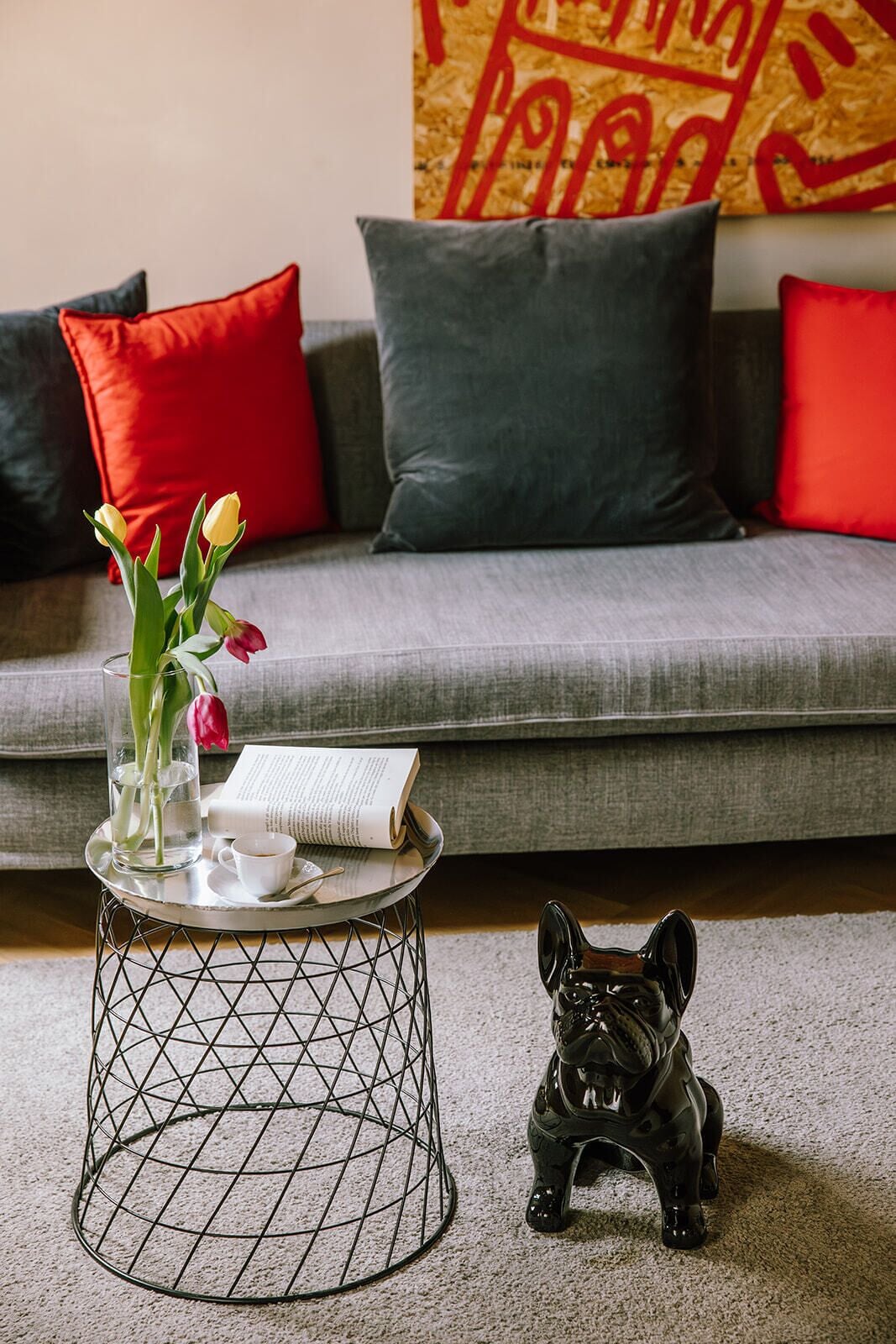 Een woonkamer met een grijze bank, rode en grijze kussens, een kleine ronde tafel met een glazen vaas met bloemen, een boek en een keramieken hondbeeld.
