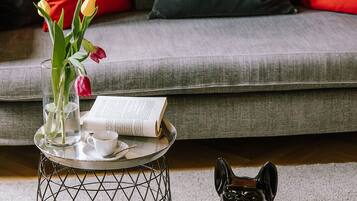 Een woonkamer met een grijze bank, rode en grijze kussens, een kleine ronde tafel met een glazen vaas met bloemen, een boek en een keramieken hondbeeld.