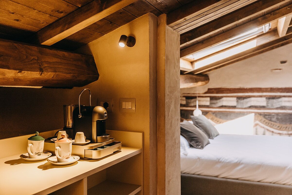 A cozy room with a wooden ceiling, a bed with pillows, a bedside table with a coffee maker, cups, and a kettle.