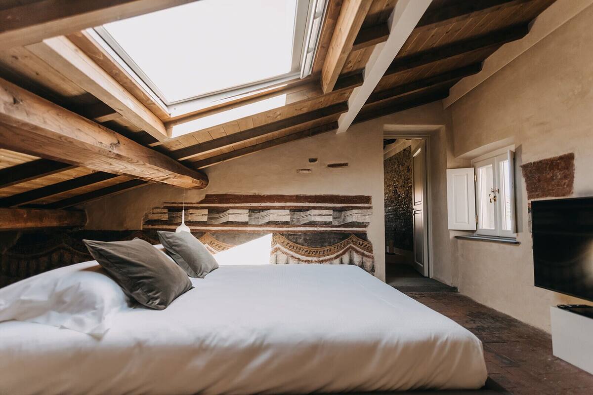 A bedroom with a skylight, wooden beams, a bed with pillows, a TV, and a stone wall.