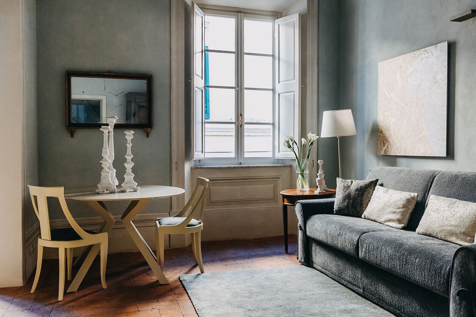 Een kamer met een grijze bank, een ronde tafel met stoelen, een staande lamp en een schilderij aan de muur.