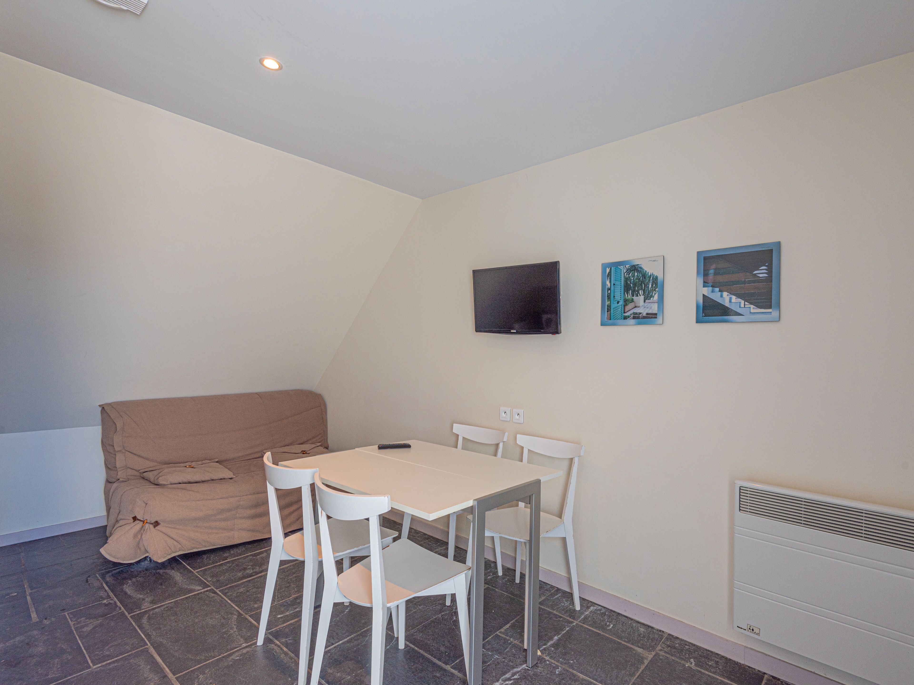 Une petite pièce bien éclairée, avec une seule table et quatre chaises, un téléviseur à écran plat fixé au mur et un radiateur en dessous.