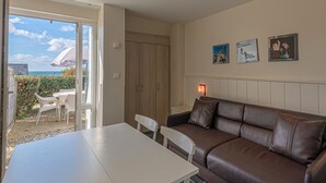A room with a brown leather sofa, a white table, and chairs, a door leading to a balcony with a view of the sea, and three framed pictures on the wall.