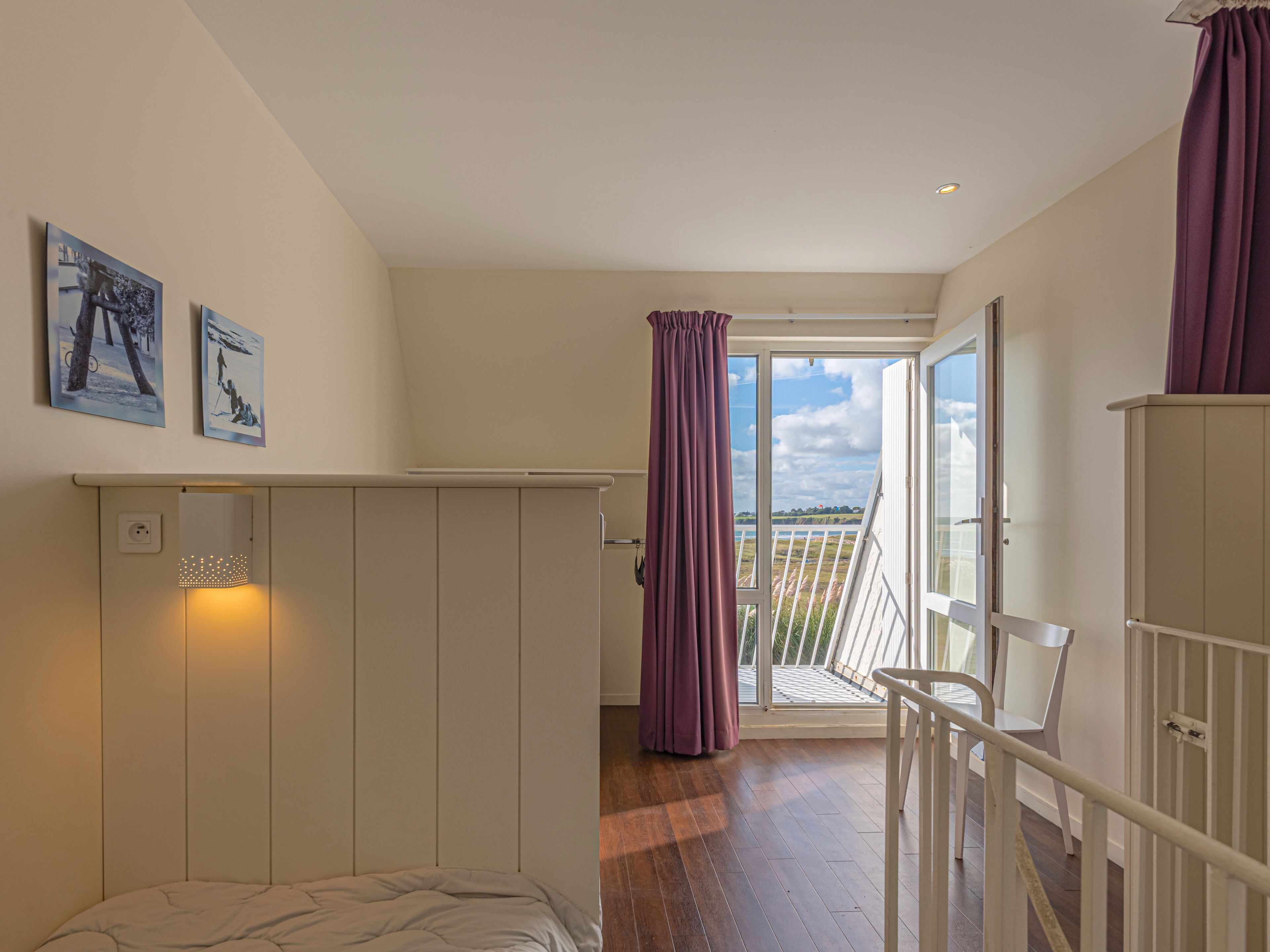 Une chambre à coucher avec un lit, une fenêtre avec des rideaux, un balcon avec une balustrade et deux tableaux accrochés au mur.