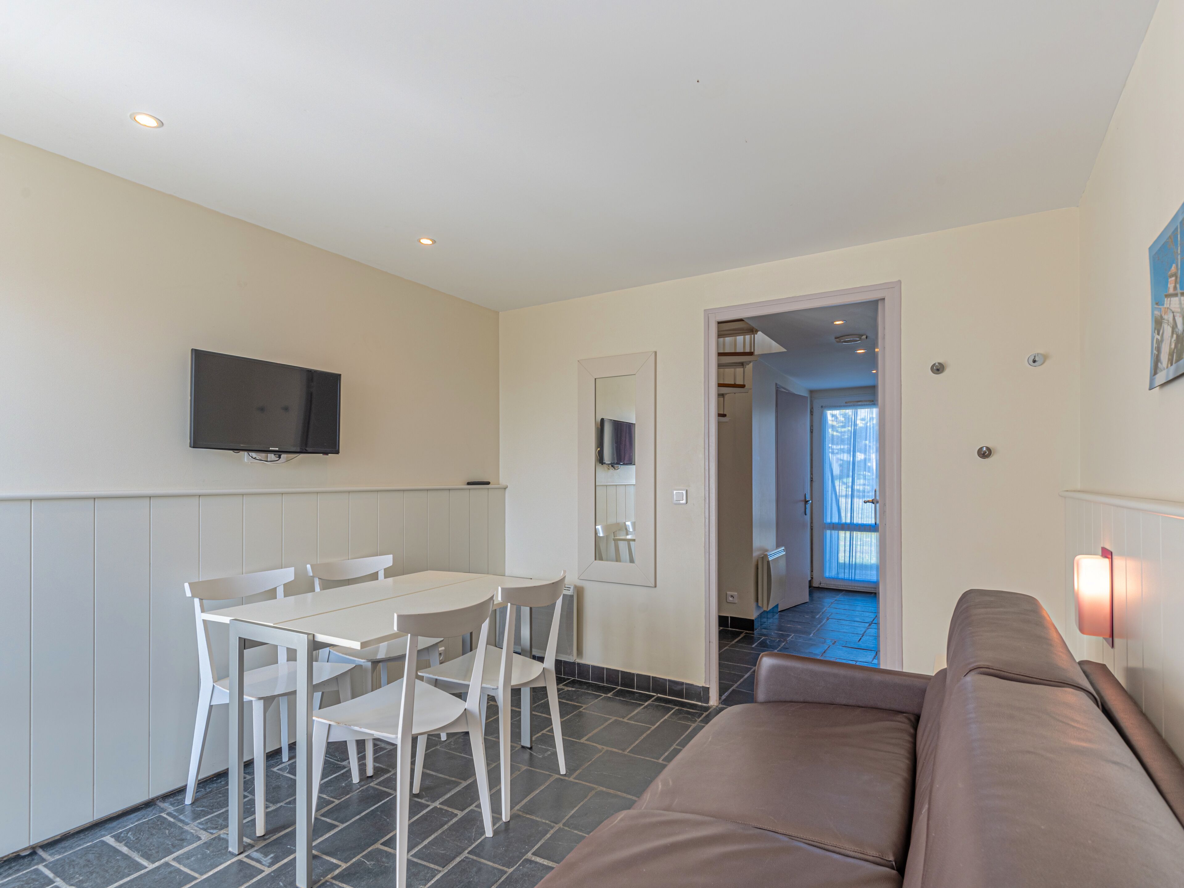 Een moderne woonkamer met een bruine bank, een eettafel met witte stoelen, een flatscreen-tv aan de muur en een deur naar een andere kamer.