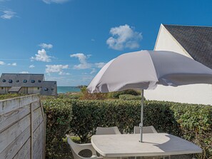 A patio with a table and chairs, an umbrella, and a view of the sea and buildings. - Vacancéole - Résidence Sud Océan (Guidel)