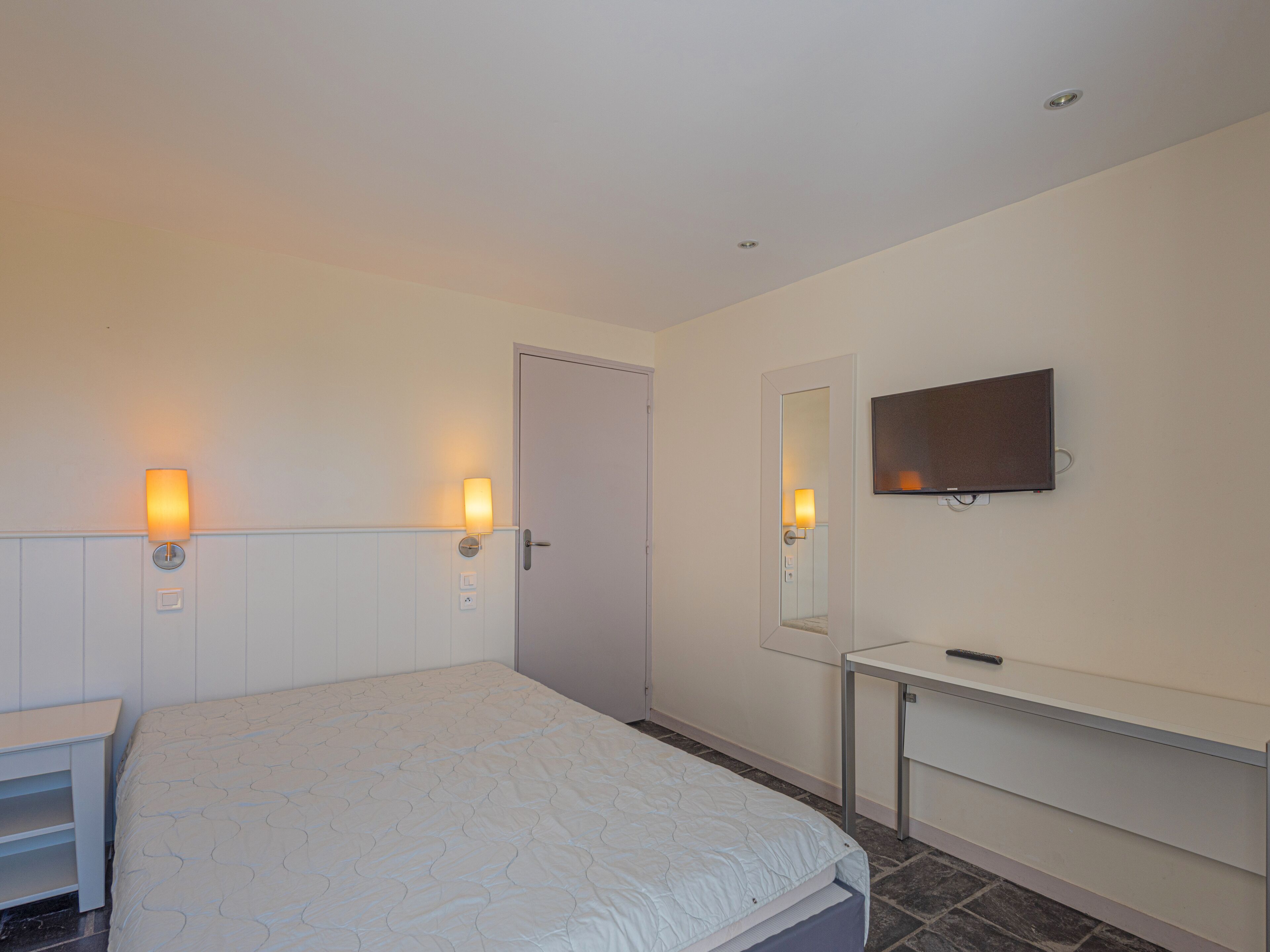 A hotel room with a bed, bedside lamps, a TV mounted on the wall, a door, and a small table.