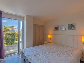 Quarto com cama, guarda-roupa, mesa de cabeceira pequena e janela com vista para o verde.