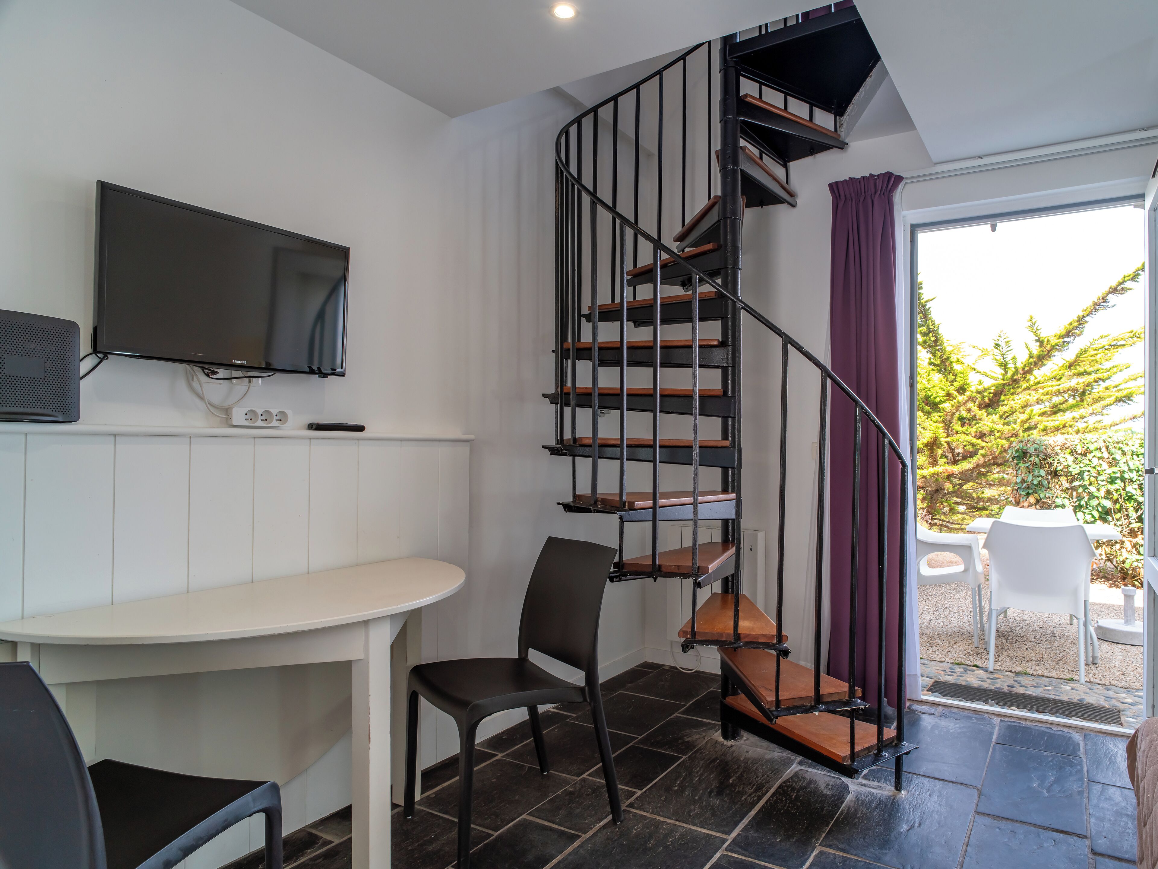 A modern room with a spiral staircase, a flat-screen TV, a dining area, and a view of the outdoors.
