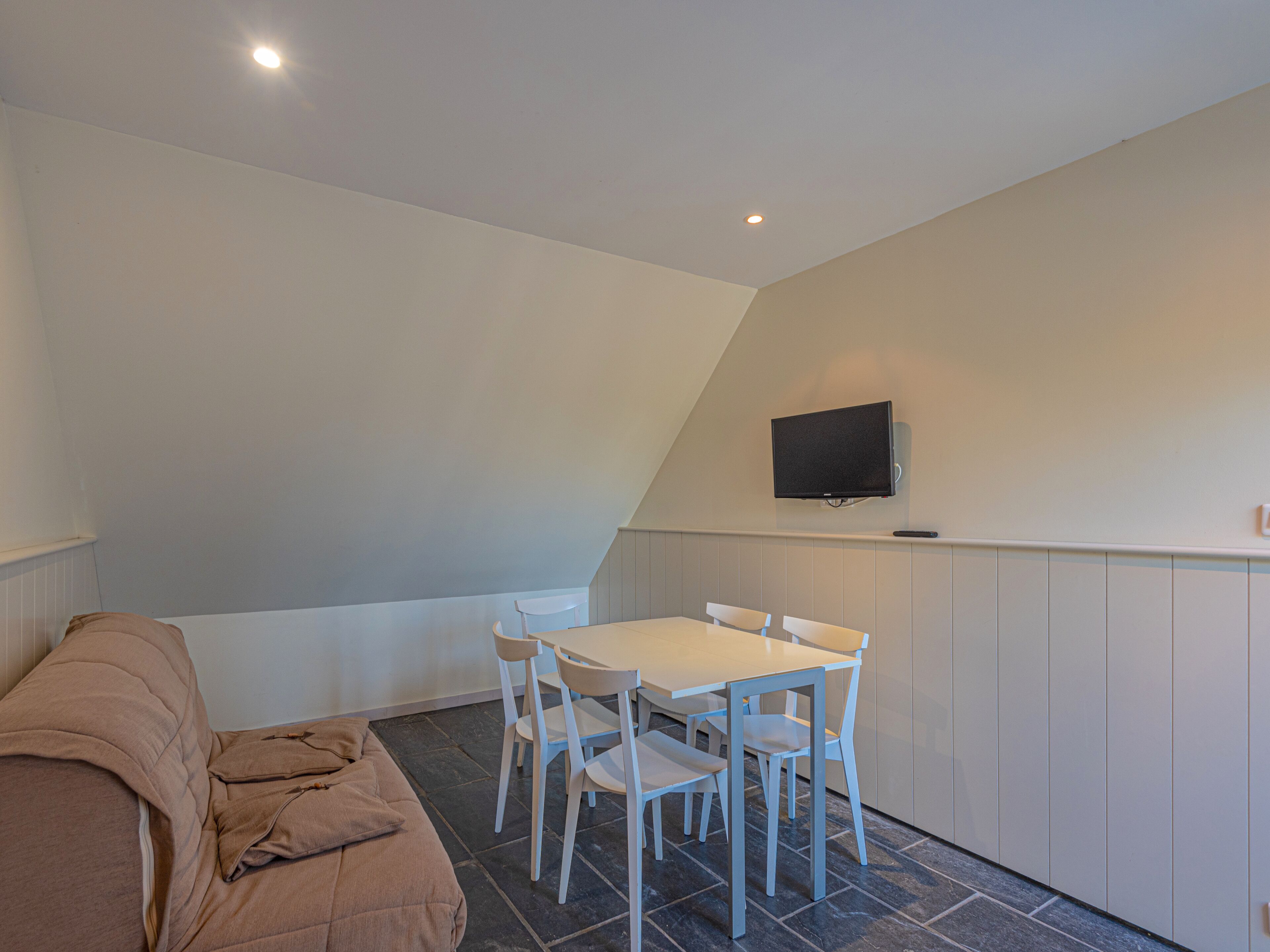 A small, modern room with a white table and chairs, a flat-screen TV mounted on the wall, and a brown sofa.