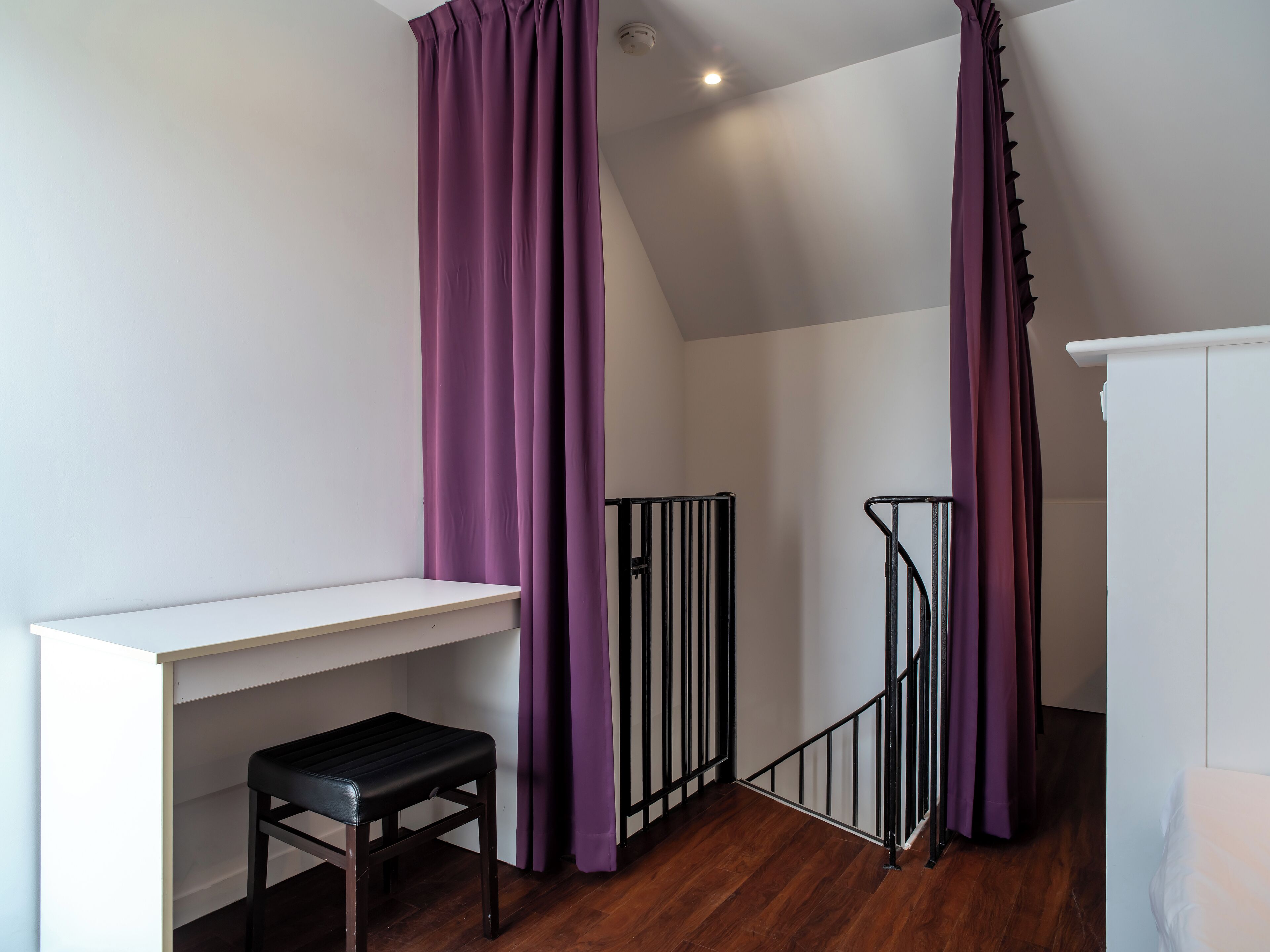 Une pièce avec un bureau blanc, un tabouret noir, des rideaux violets et une armoire blanche.