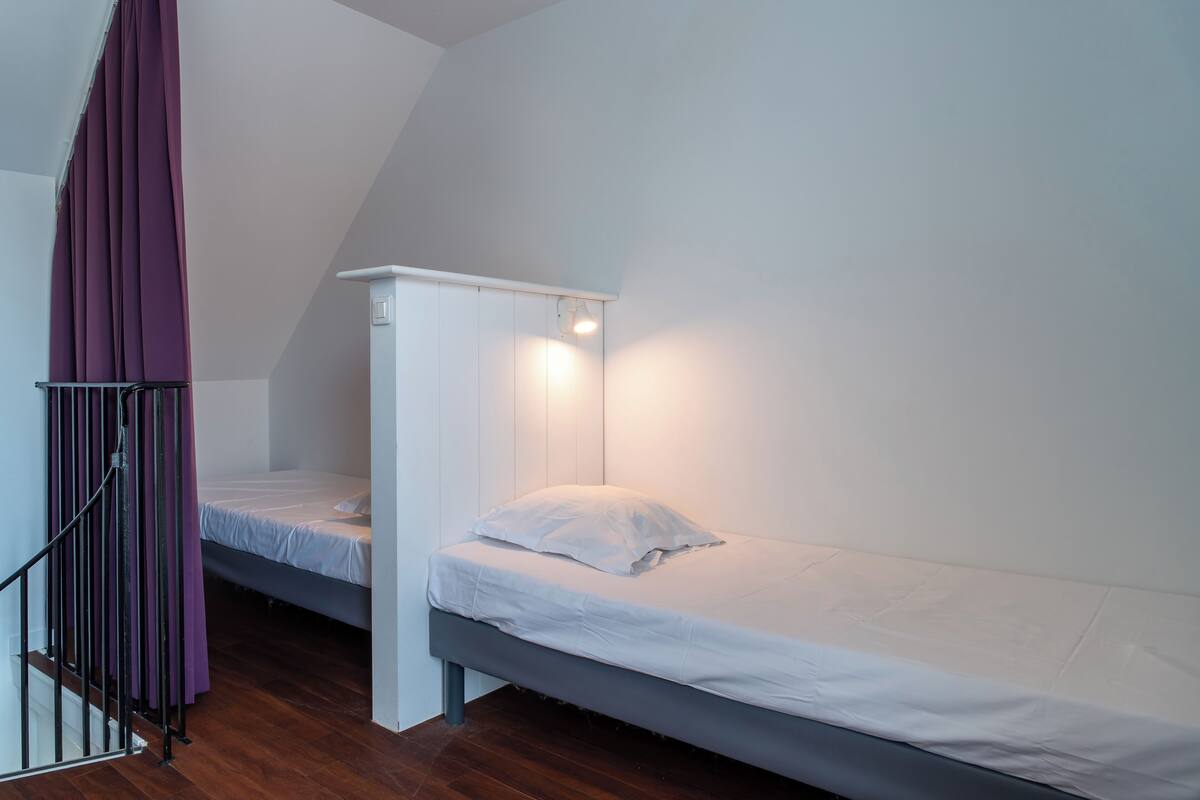 Una habitación pequeña con cama individual, techo inclinado, pared blanca y suelo de madera.
