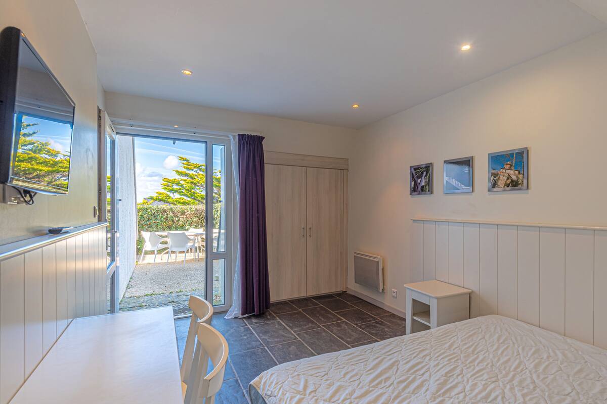 Una habitación de hotel moderna con una cama, un escritorio y una silla. Hay una puerta corredera que da a un espacio exterior con sillas y una mesa.