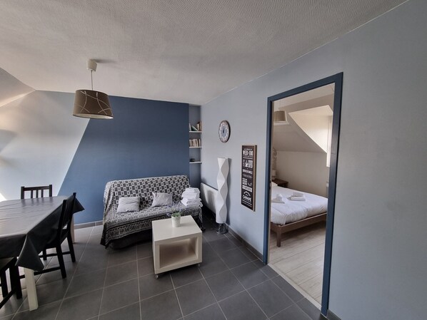 Una habitación moderna y compacta con una pared azul de acento, un sofá, una pequeña mesa de centro, una mesa de comedor con sillas, y una cama en una habitación contigua.