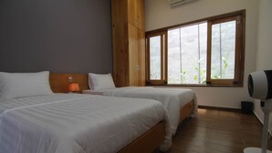 Une chambre d’hôtel avec deux lits, un plancher en bois, une grande fenêtre donnant sur des plantes et un ventilateur de plafond.