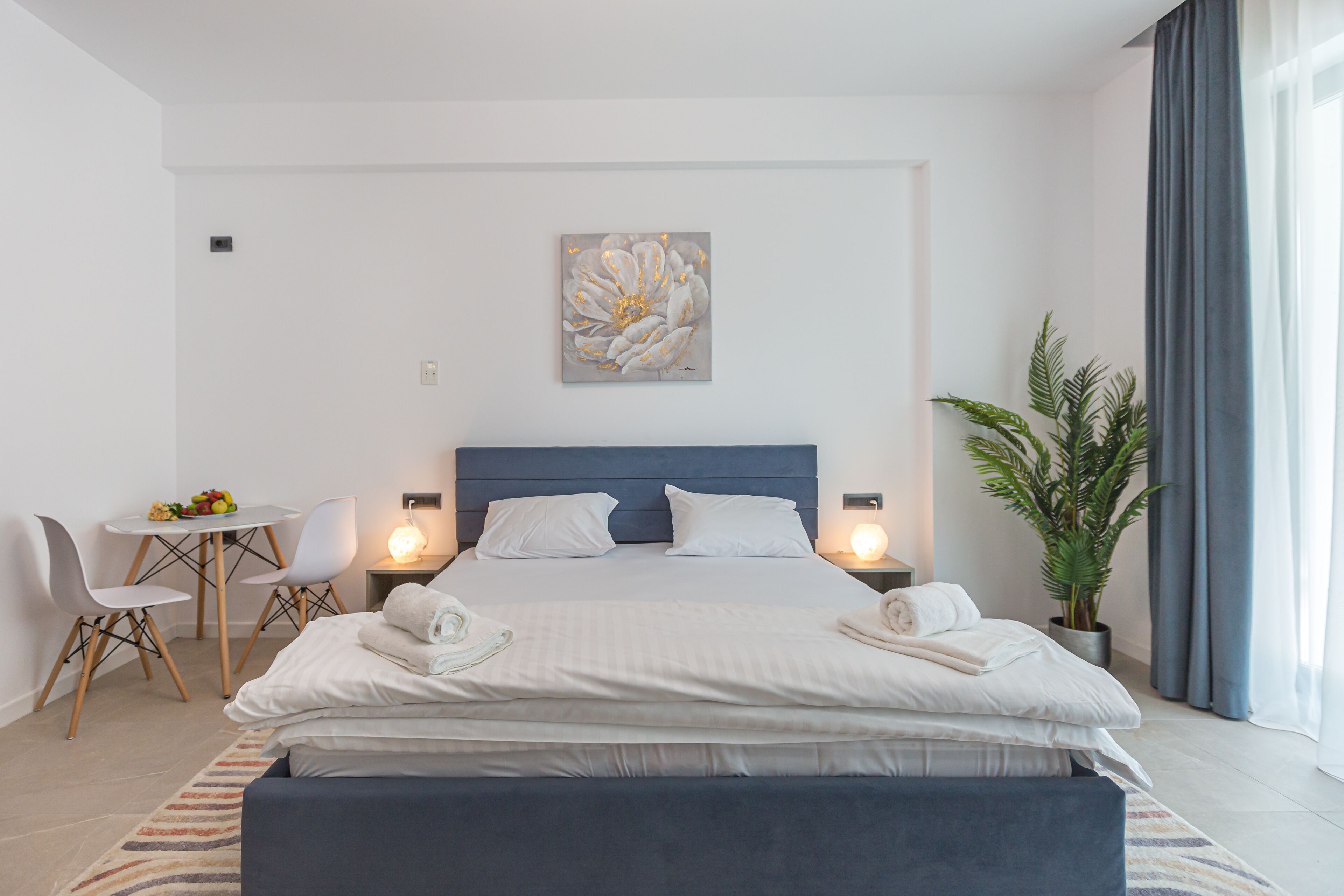 A modern bedroom with a large bed, bedside tables, a desk with chairs, a potted plant, and a wall-mounted artwork.