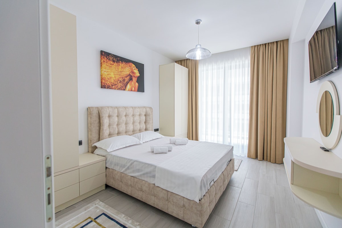 A modern hotel room with a large bed, a wardrobe, a vanity with a mirror, and a painting on the wall.