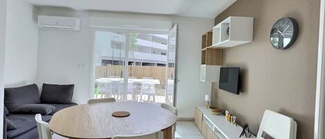 Un salon moderne avec une table ronde en bois, des chaises blanches, un canapé gris et un téléviseur fixé au mur.