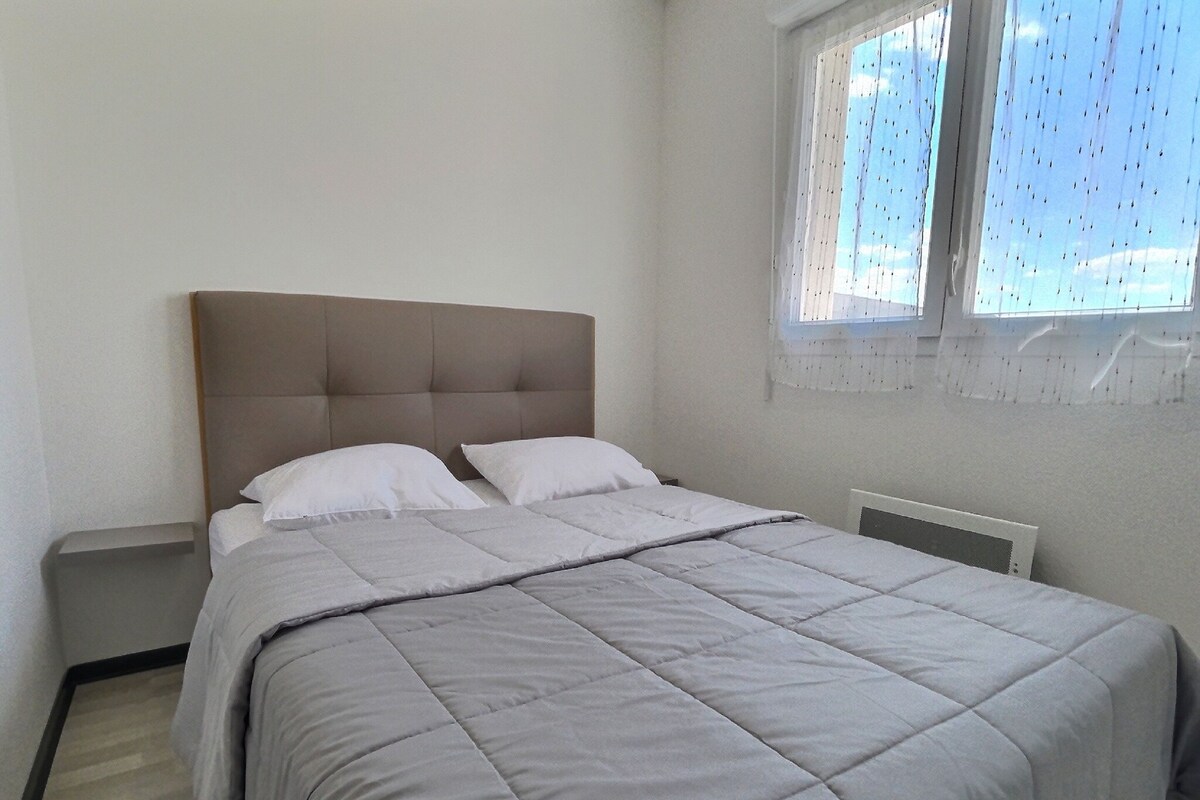 Una cama bien hecha con colcha gris y almohadas blancas en una habitación sencilla con ventana y radiador.