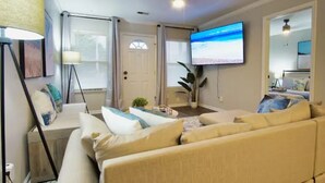 Ett vardagsrum med en beige soffa, ett soffbord, en golvlampa och en tv som hänger på väggen.