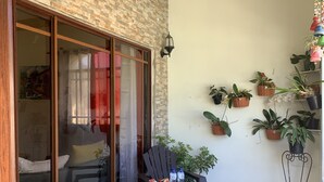 A patio with two wooden chairs, a small table with snacks, and potted plants on a balcony with a brick wall and sliding glass doors.