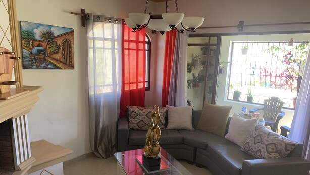 A living room with a glass coffee table, a sofa, and a couch. There is a painting on the wall and a window with curtains.