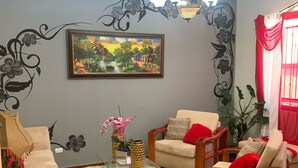 A living room with a wooden floor, a glass coffee table, two beige armchairs with red cushions, a framed painting on the wall, and a chandelier.