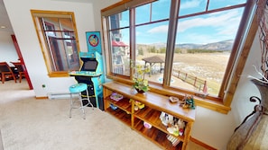 A room with a large window, a wooden desk, a chair, and a shelf with various items.