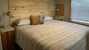 Un dormitorio con cabecera de madera, cubrecama a rayas, dos almohadas, una lámpara de noche y una ventana con persianas.