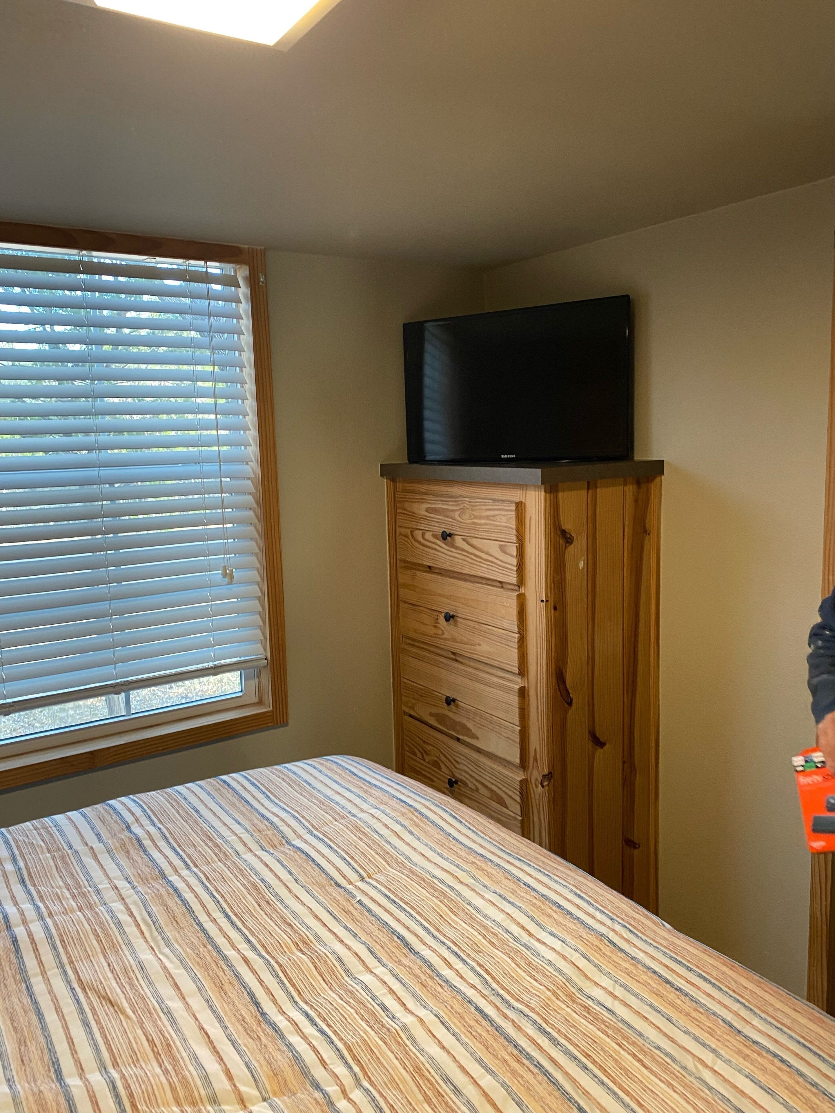 Une chambre à coucher avec un lit, une commode en bois, un téléviseur posé dessus et une fenêtre avec des stores.