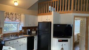 Una cocina compacta con armarios blancos, un refrigerador negro, una cafetera y un televisor.