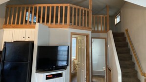 Interior de una vivienda de varios niveles con una escalera de madera, una zona de cocina con un refrigerador negro y un televisor montado en la pared.