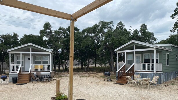 Una terraza de madera con una parrilla y sillas, dos cabañas con porches, y suelo de grava.