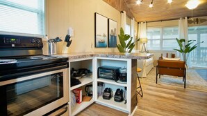 A modern kitchen with a built-in oven, a coffee machine, and a toaster. There is a white cabinet with shelves, a plant, and a chair. - Serenity Sol ※ Surfside Charmer Near Beach & Jetty (Surfside Beach)