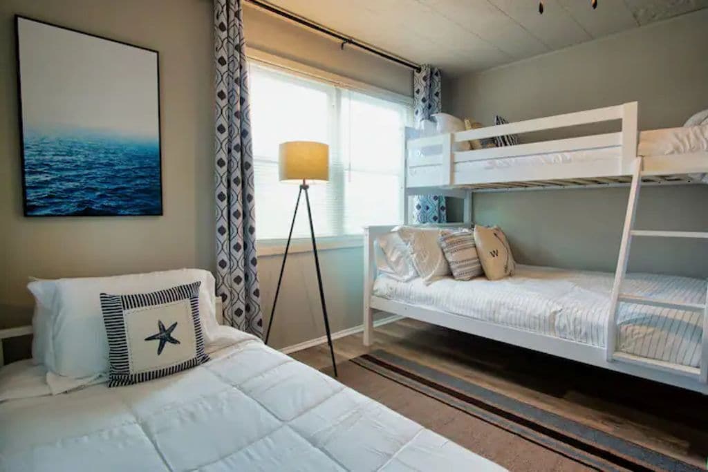A hotel room with a bunk bed, a bed with a star-shaped pillow, a floor lamp, and a painting of the ocean.