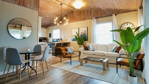 A modern living room with a wooden ceiling, a dining area with chairs and a table, a sofa with cushions, and a large mirror on the wall. - Serenity Sol ※ Surfside Charmer Near Beach & Jetty (Surfside Beach)