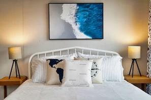 Ein Hotelzimmer mit einem Bett, zwei Nachttischlampen, einem Kopfkissen mit Weltkarte, einem Wecker-Kissen und einem gerahmten Kunstwerk mit einem Wellenmotiv über dem Bett.