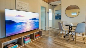 A modern room with a large flat-screen TV displaying a beach scene, a wooden TV stand, a dining area with a round table and chairs, and a 'Welcome to our Beach House' sign. - Serenity Sol ※ Surfside Charmer Near Beach & Jetty (Surfside Beach)