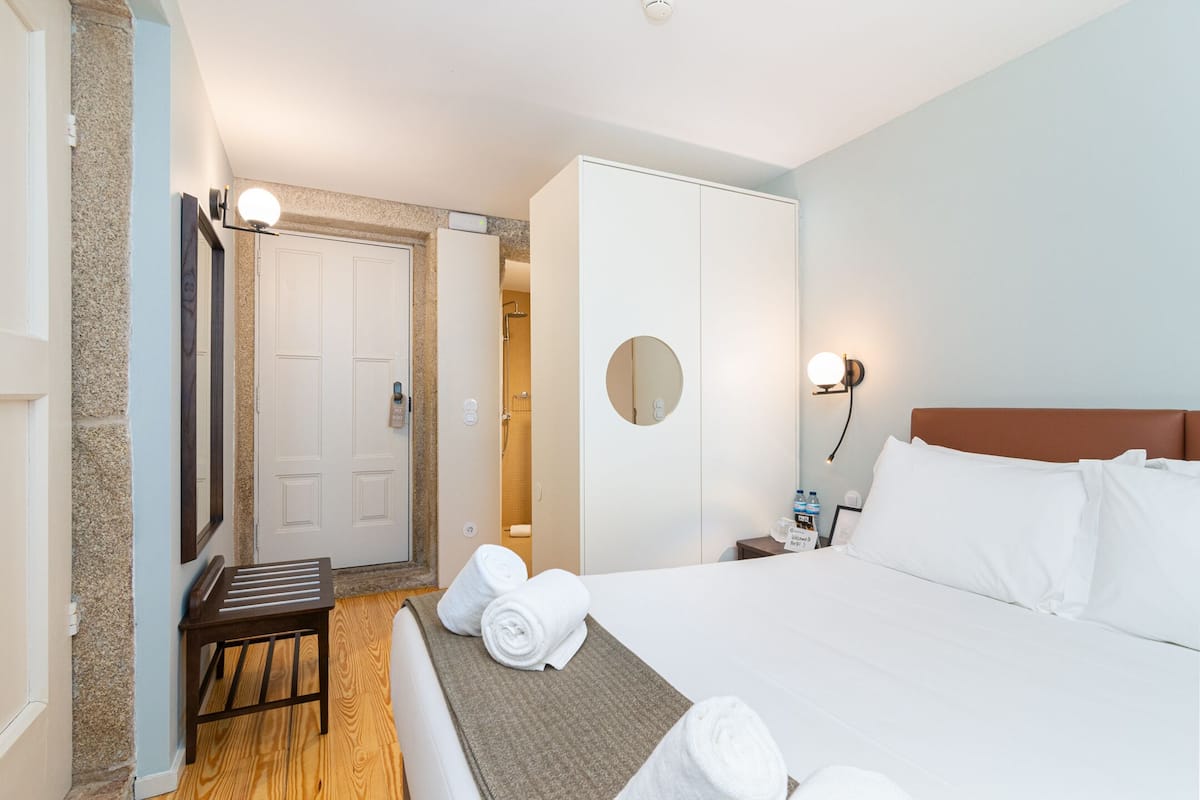 A hotel room with a bed, a bedside table, a mirror, a wardrobe, and a small stool.