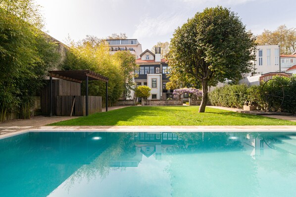 A swimming pool with a wooden deck and a pergola, surrounded by a well-maintained lawn and trees.