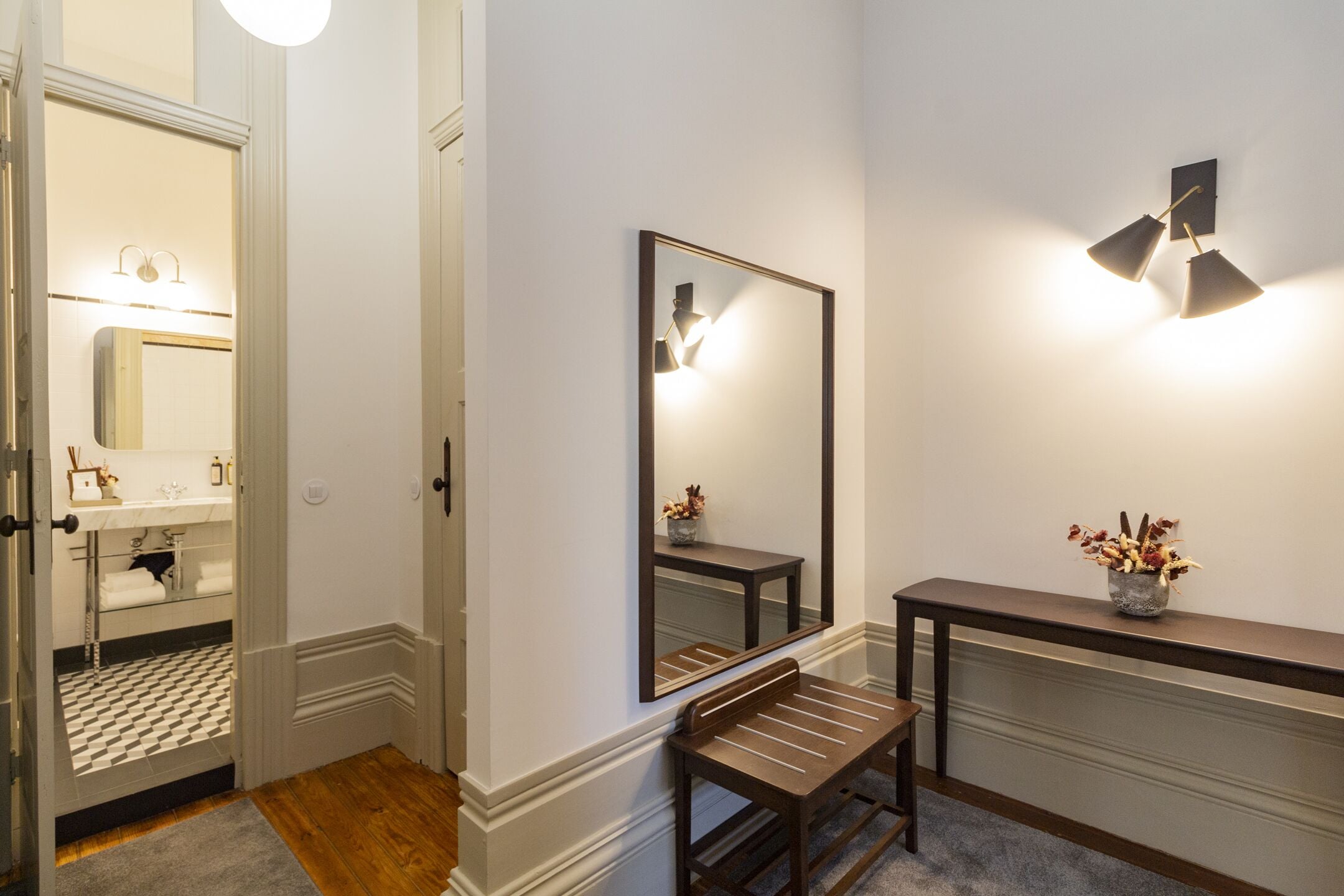 A hallway with a mirror, a bench, a vase with flowers, and a wall-mounted light fixture.