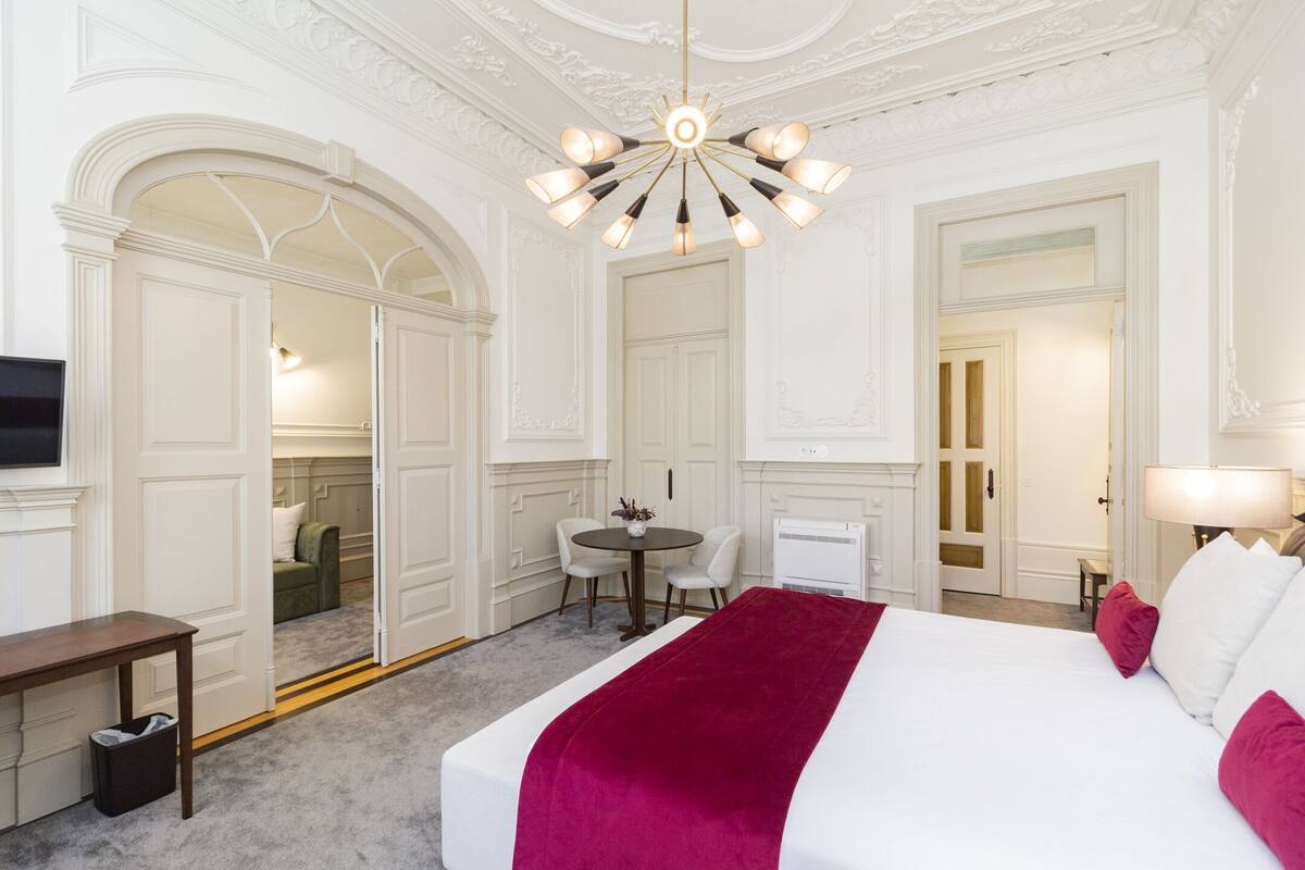 A hotel room with a large bed, a desk, a chair, a table, and a chandelier.