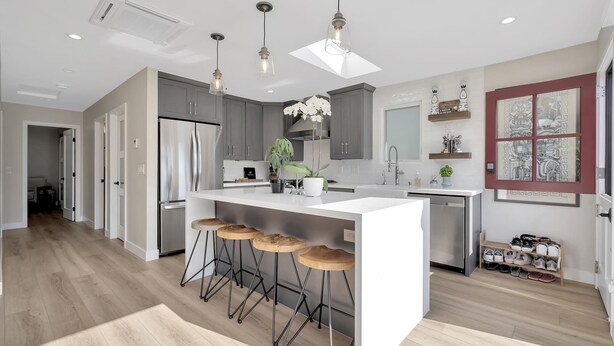 Una cocina moderna con una isla central, taburetes de bar, iluminación colgante y lavavajillas integrado.