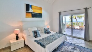 A bedroom with a bed, nightstands, a large painting, and a view of the outdoors through a sliding glass door. - Callisto (2/3 Rooms) Luxury villa (Les Terres Basses)