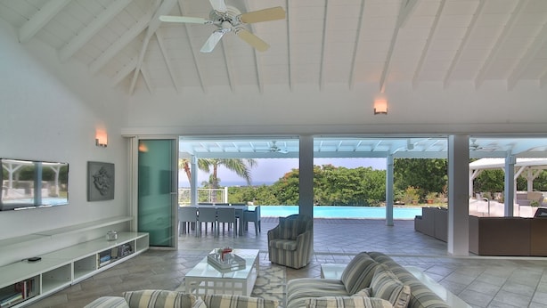 A spacious living area with a large sofa, a coffee table, and a TV mounted on the wall. There is a sliding glass door leading to an outdoor patio with a pool and a view of the ocean. - Callisto (2/3 Rooms) Luxury villa (Les Terres Basses)