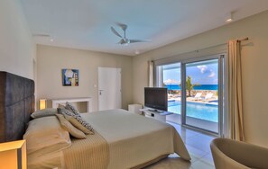 Ein Schlafzimmer mit Bett, Fernseher, Deckenventilator, einem Gemälde und Blick auf Pool und Strand.