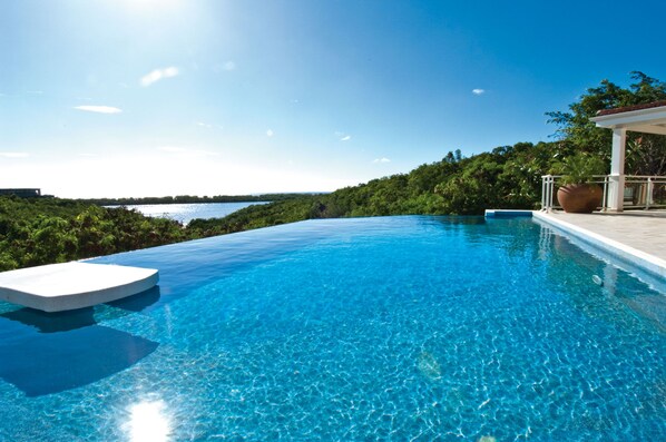 Une magnifique piscine à débordement d’un bleu profond, bordée de blanc, entourée d’une végétation luxuriante et offrant une vue sur la mer.