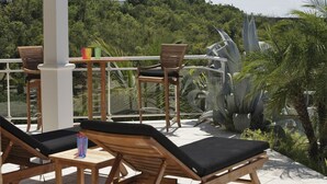 A terrace with wooden lounge chairs and a table, surrounded by tropical plants and a view of a lush hillside.